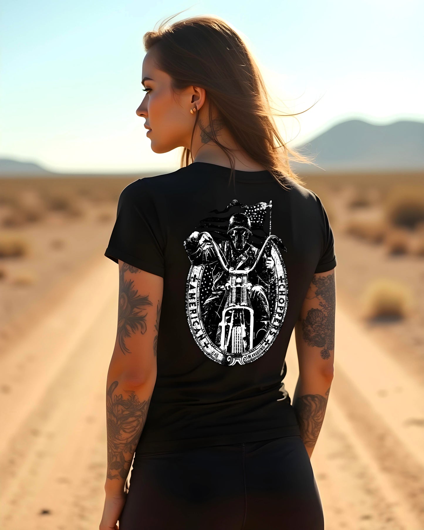 A tattooed woman stands in the desert wearing a black T-shirt with a large backprint of a skull-faced biker on a chopper, backed by a waving American flag.